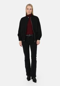 Giacca bomber nera con zip frontale e due tasche anteriori, indossata sopra un dolcevita bordeaux e abbinata a jeans neri svasati e stivaletti.