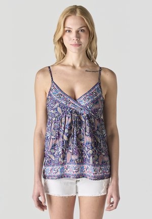 Mujer rubia con blusa sin mangas de tirantes finos con estampado de cachemira azul y pantalones cortos blancos con flecos, posando frente a un fondo liso.