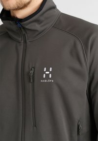 Mann, der eine dunkelgraue Zip-Jacke mit hohem Kragen und gepolsterter Brusttasche trägt, mit weißem Haglöfs-Logo auf der linken Brust.