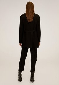 Sort skræddersyet blazer med en knyttet talje, parret med matchende bukser og spidse sorte hæle, glat stof og elegant design.