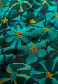 Tyrkysová látka s překrývajícími se temně modrými květinovými vzory, každý květ je uprostřed s malými hořčičně žlutými okvětními lístky, což ukazuje jemné záhyby a texturu.
