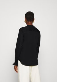 Camicia nera a maniche lunghe con una texture liscia, chiusura a bottoni sul retro e polsini arricciati, abbinata a pantaloni ampi color crema.