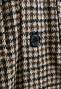 Tessuto a quadri marrone e beige con una texture pelosa, dotato di un bottone nero con dettagli in rilievo. Dettaglio ravvicinato del design.