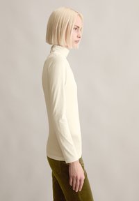 Cremefarbenes Langarm-Turtleneck-Oberteil mit taillierter Passform, hergestellt aus einem glatten, weichen Material. Kombiniert mit olivgrünen Cordhosen.