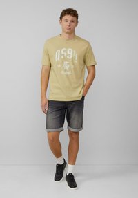 Lichtgele t-shirt met witte grafische print, gecombineerd met donkergrijze denim short met opgerolde zoom. Zwarte sneakers met witte zolen.