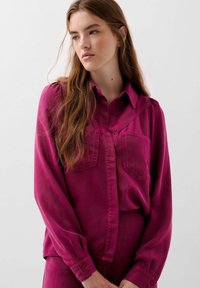 Camicia magenta con bottoni, due tasche sul petto, maniche lunghe e colletto. Il tessuto ha una consistenza liscia, con dettagli di cucitura sottili.