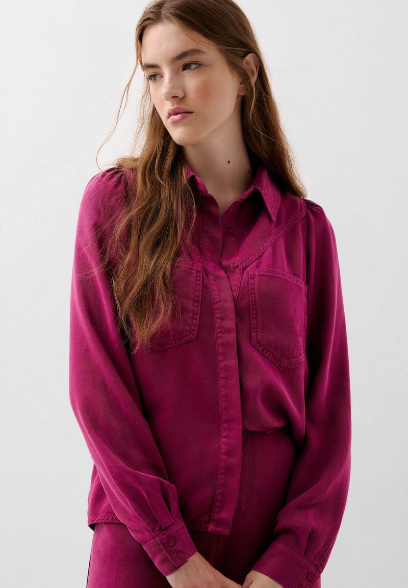 Camicia magenta con bottoni, due tasche sul petto, maniche lunghe e colletto. Il tessuto ha una consistenza liscia, con dettagli di cucitura sottili.