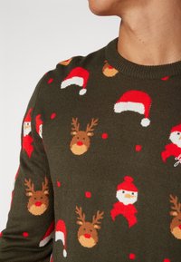 Grön stickad tröja med ett festligt mönster av röda tomteluvor, renhuvuden och snöflingor, med en texturerad yta och rundad halsringning.