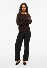 Brūna teksturēta džemperis ar ribotām piedurknēm, kombinēts ar melnām brīvām biksēm un norādītām melnām kurpēm ar augstu papēdi. Modelis stāv ar sakrustotām rokām.