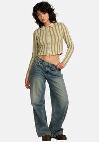 Camisa de manga larga recortada en beige con rayas verticales verdes y amarillas; combinada con vaqueros azules holgados y botas marrones.