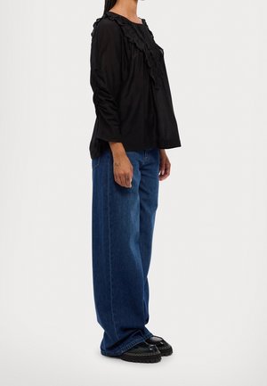 Schwarzes Langarmoberteil mit Rüschensaum, kombiniert mit locker sitzenden blauen Jeans. Die Schuhe sind schwarze Plateau-Schuhe.
