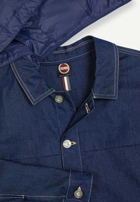 Dunkelblaues Jeanshemd mit beigen Nähten, Metall-Druckknöpfen und einer teilweise sichtbaren marineblauen gesteppte Jacke oberhalb des Kragens.