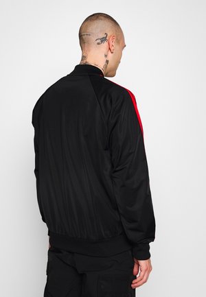 Mężczyzna z tatuażami, ubrany w czarną kurtkę z czerwonymi paskami na rękawach, stoi odwrócony tyłem na tle jednolitego jasnego tła.