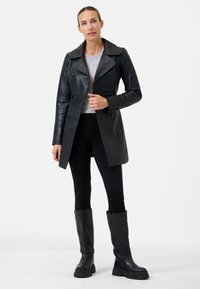 Black leather trench coat with a wide collar and double-breasted buttons, worn over a gray sweater and paired with black leggings and tall boots.