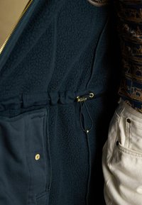 Sötét tengerészkék gyapjú dzseki belső része húzózsinórral, cipzáras zsebbel és aranyszínű kiegészítőkkel. Jellemzője a texturált gyapjú anyag.