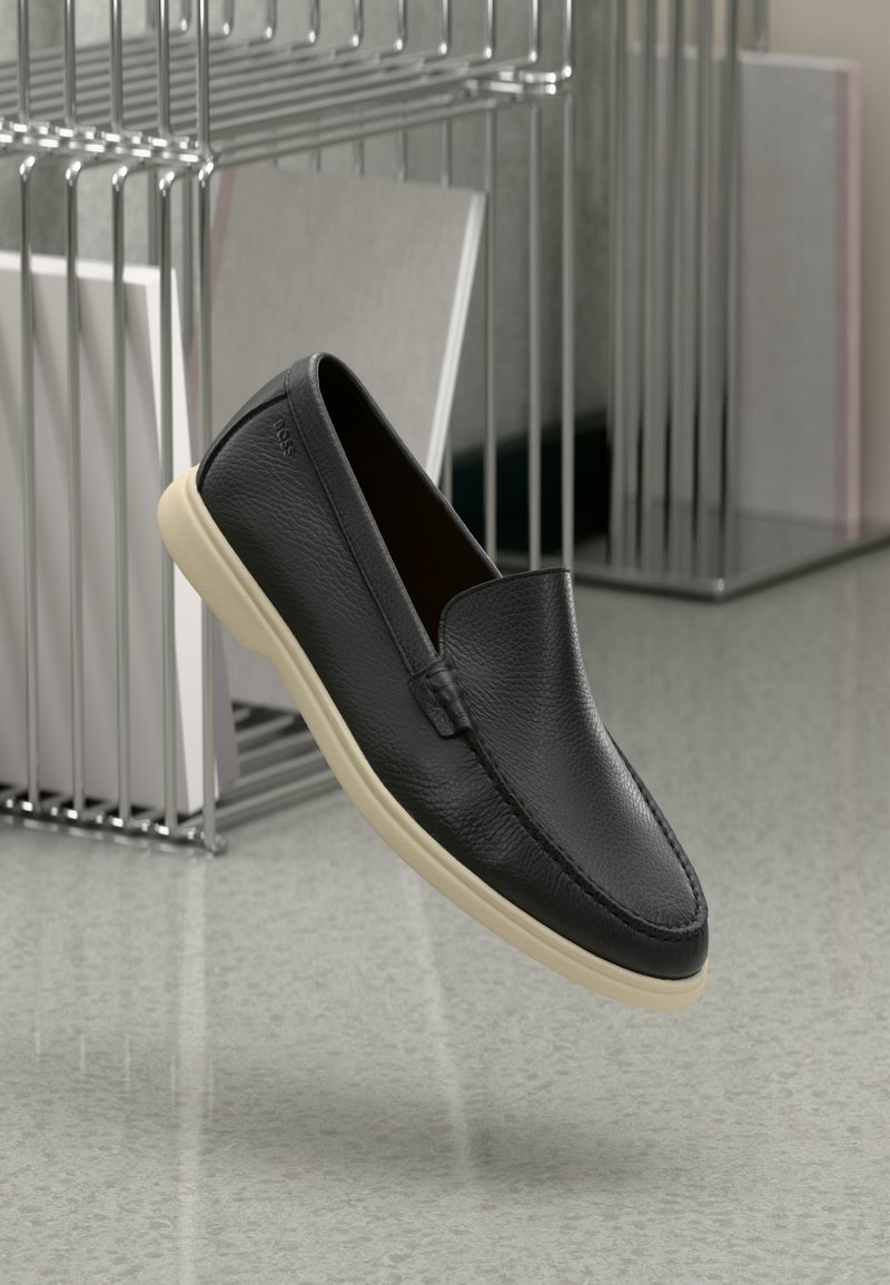 Schwarzer Loafer aus Leder zum Hineinschlüpfen mit beiger Sohle, schwebend in der Luft nahe einem Metall-Draht-Magazinhalter auf einem Terrazzo-Boden.