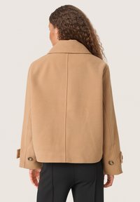 Krátky kabát v camel farbe vyrobený z jemného materiálu, s klasickým golierom, širokými rukávmi a zaoblenými gombíkmi na manžetách.