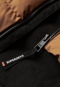 Brązowa puchowa kurtka z czarnym futrem wewnętrznym i zamkiem błyskawicznym. Posiada markową czarną metkę z napisem "SUPERDRY" przy uchwycie zamka.