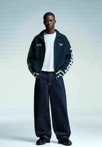 Jeune homme portant une veste zippée bleu marine avec du texte blanc, une chemise blanche, un pantalon large foncé et des chaussures noires se tenant contre un fond clair.