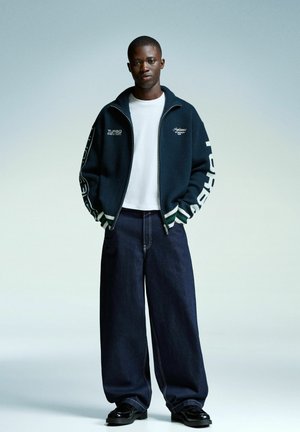 Unge mand iført en navy zip-up jakke med hvid tekst, hvid skjorte, brede mørke jeans og sorte sko, der står foran en lys baggrund.