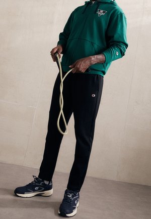 Persona con sudadera con capucha verde, pantalones deportivos negros y zapatillas oscuras sosteniendo una cuerda para saltar beige anudada frente a una pared de azulejos.