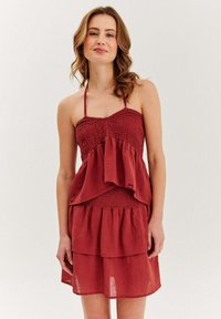 Žena v bezrúrkových, vrstvených červených šatách na ramienkach, stojaca proti jednoduchému svetlému pozadiu, s jemným úsmevom.