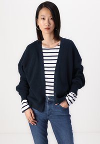 Granatowy sweter dziany z szerokimi rękawami, otwarty z przodu i o prążkowanej strukturze, założony na biało-granatową, w paski koszulę z długimi rękawami.