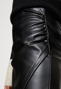 Černá kožená sukně s nařasenými detaily a diagonálními švy, vykazující hladkou texturu a jemný lesk.