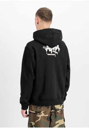 Man wearing black hoodie with white "ANIMA Industries" logo on back, paired with green and brown camouflage cargo pants, facing away.