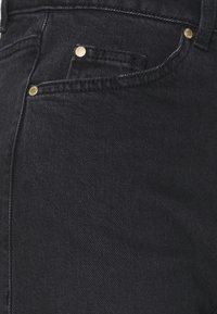 Pantalones de mezclilla negra con textura suave, con detalles de herrajes dorados y un diseño clásico de cinco bolsillos. Primer plano de la sección frontal.