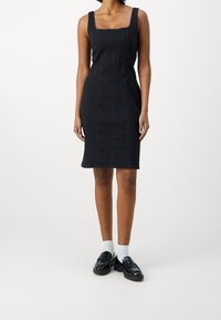 Musta farkkutunika, jossa on neliskulmainen pääntie, tiukka yläosa ja kynähame. Yhdistetty mustien loafer-kengien ja valkoisten sukkien kanssa. Sujuva rakenne.