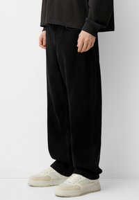 Črne prevelike kavbojke iz denima z stranskimi žepi, v kombinaciji s kremnimi športnimi copati. Gladka tekstura in sproščen kroj poudarjata ležeren dizajn.