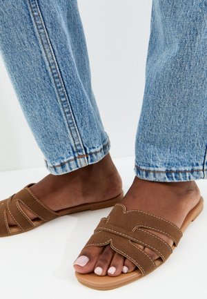 Pies calzados con sandalias sin cordones de ante marrón combinadas con vaqueros de mezclilla azul claro, mostrados sobre un fondo blanco liso.