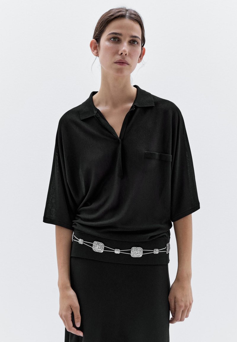 Sort kortærmet polo-shirt med krave, med en brystlomme og et bælte med sølvaccenter. Blødt, struktureret stof. Minimalistisk design.