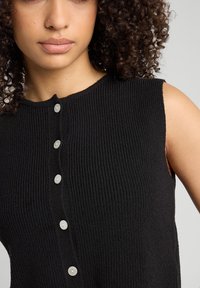 Mustat ribbitetut hihattomat paidat, joissa on pyöreä kaula-aukko ja viisi valkoista nappia edessä. Teksturoitu kangas, jossa on kevyt venyvyys.