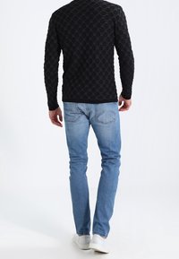 Un pull noir à manches longues avec un motif circulaire texturé, associé à un jean slim bleu clair et des baskets blanches, vu de dos.