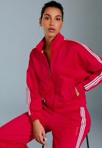 Compleu roșu dintr-un material neted; include o jachetă cu fermoar și dungi albe pe mâneci, precum și pantaloni asortați. Detalii minime de branding vizibile.