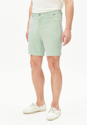 Homme portant un short vert clair jusqu'aux genoux, un t-shirt blanc et des chaussures bateau blanches, debout devant un fond blanc uni.