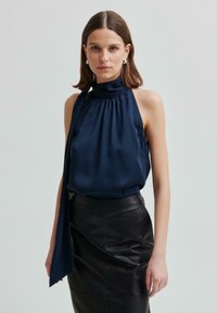 Top halter in seta navy con scollatura arricciata e lunga cravatta drappeggiata, abbinato a una gonna a tubino in pelle nera. Texture liscia, design classico.