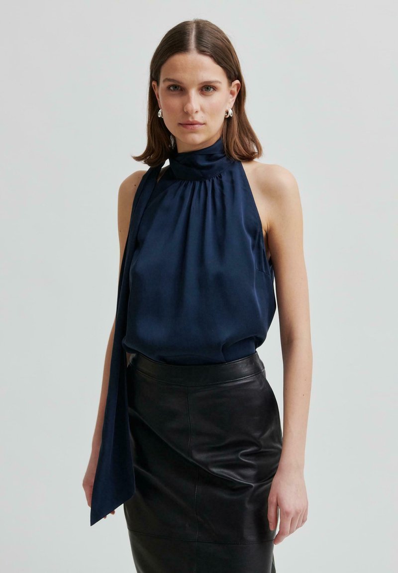 Top halter in seta navy con scollatura arricciata e lunga cravatta drappeggiata, abbinato a una gonna a tubino in pelle nera. Texture liscia, design classico.