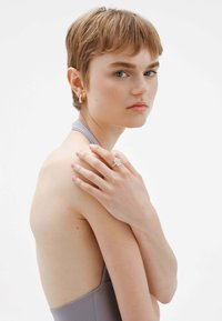 Anillo de oro con una gema cuadrada y transparente. El modelo lleva una prenda sin mangas de color gris, que muestra el hombro y el cuello. Pendientes de aro de oro.