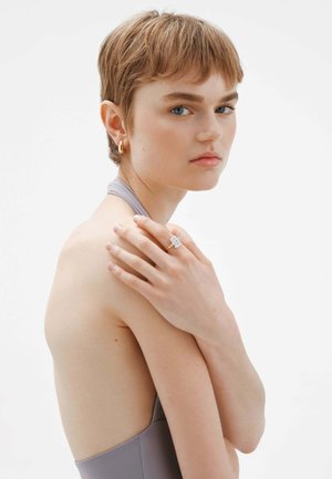 Anillo de oro con una gema cuadrada y transparente. El modelo lleva una prenda sin mangas de color gris, que muestra el hombro y el cuello. Pendientes de aro de oro.