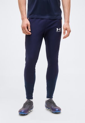 Mężczyzna ubrany w granatowe spodnie sportowe Under Armour i czarne korki z niebieskimi akcentami stojący na jednolitym tle.