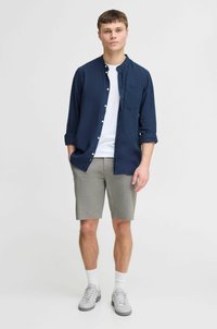 Chemise bleu marine avec un col, t-shirt blanc en dessous, shorts gris et baskets grises. Chaussettes blanches portées au-dessus de la cheville.