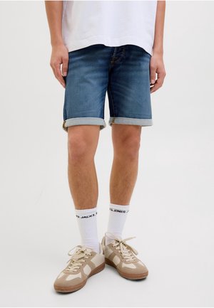 Personne portant un short en jean retroussé, des chaussettes blanches Jack & Jones et des baskets décontractées beiges, debout sur un fond clair uni.