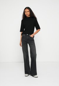 Čierny rebrovaný krátky top s krátkym rukávom v kombinácii s vysokým pásom tmavosivých rozšírených džínsov. Modelka stojí s rukami vreckách, má na sebe biele topánky.