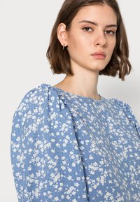 Blusa floral azul feita de tecido leve, com mangas bufantes, decote redondo e um padrão de flores brancas espalhadas.