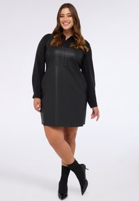 Schwarzes Kunstlederkleid mit langen Ärmeln, das einen Rüschenausschnitt und dekorative Nieten aufweist. Kombiniert mit schwarzen gerippten Socken und Absatzschuhen.