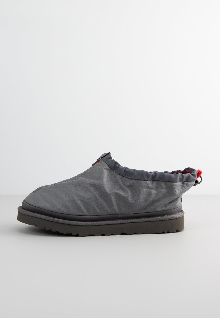 Zapato gris de tipo blando y sin cordones con un diseño curvado, cuello acolchado, suela de goma y detalle en rojo. Hecho de material ligero y resistente al agua.