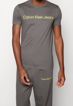 Homme portant un t-shirt et un pantalon gris assortis Calvin Klein Jeans avec un logo jaune sur la poitrine et le haut de la cuisse.